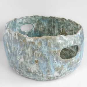 A ceramic basket with two oval cutout handles, featuring a textured glaze with mottled shades of light blue, ecru, and alabaster on a white background.